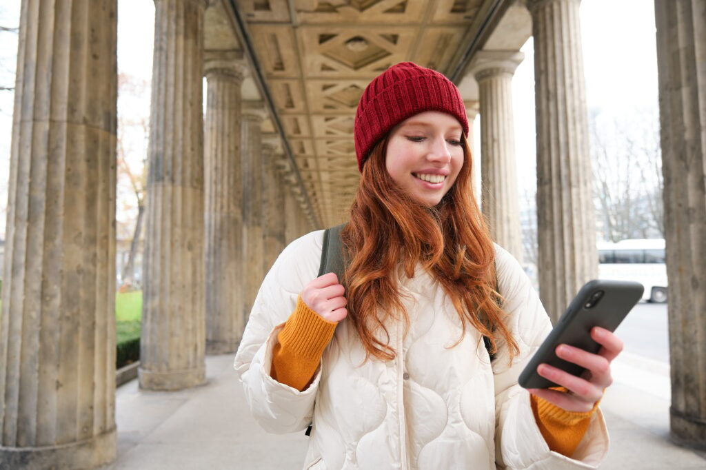 Mobile broadband and people. Smiling redhead 20s girl with backpack, uses smartphone on street